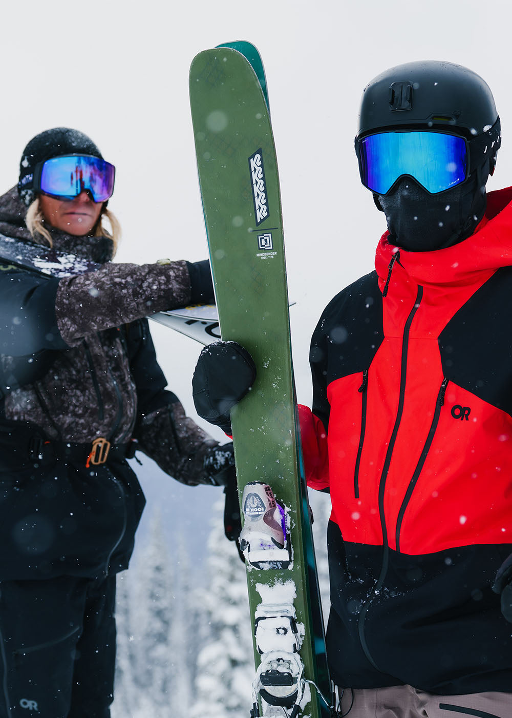 Two friends holding their skis in the foggy snow wearing Outdoor Research Men's Powderverse Jacket Ranger Green Granite Print/Black and Lingonberry/Black. 