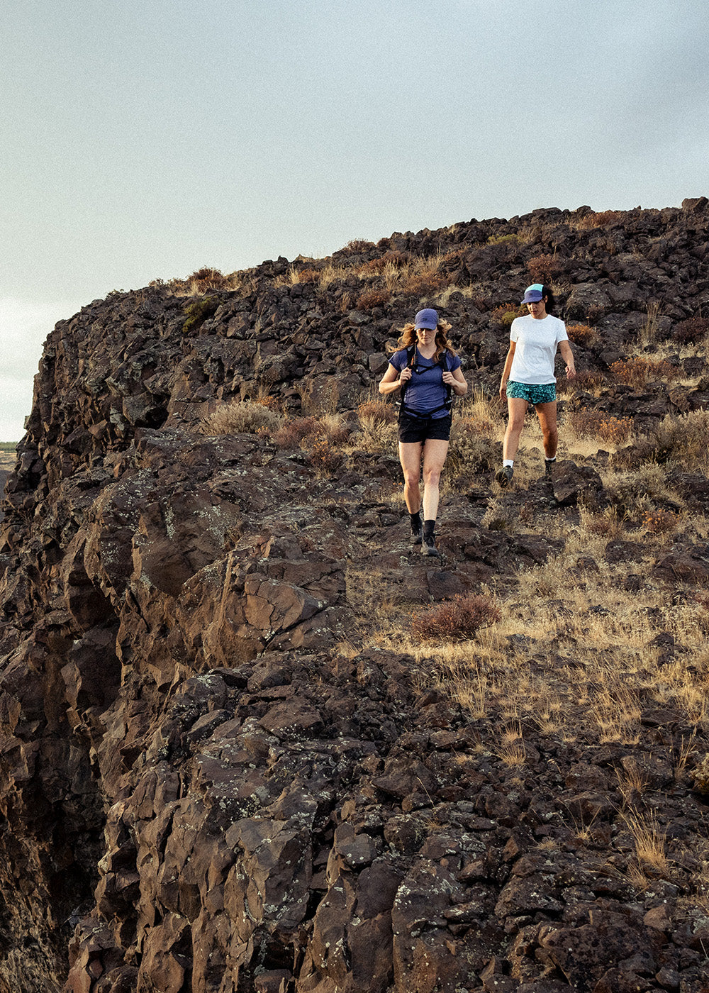 Two friends walk along the ledge of a rock cliff with a desert plateau behind them.